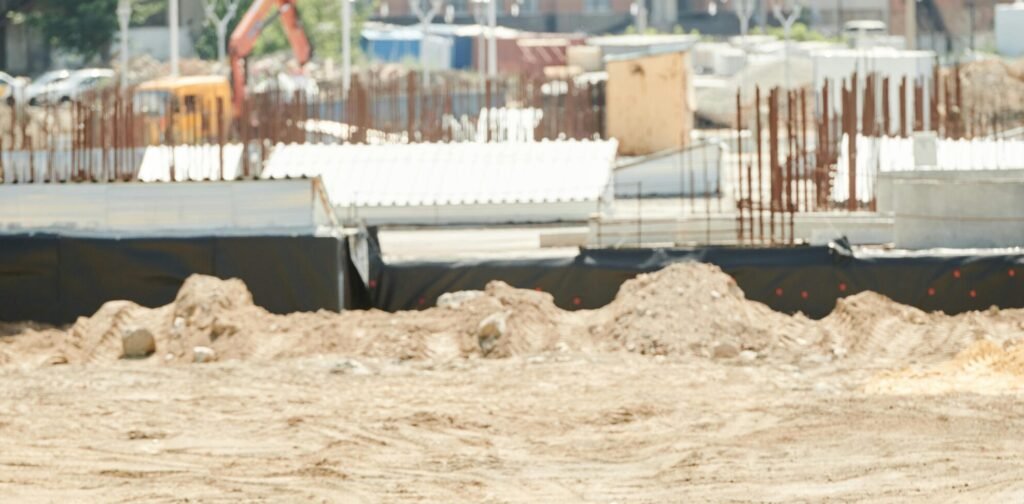 Two workers discussing plans on a sandy construction site, wearing safety gear.